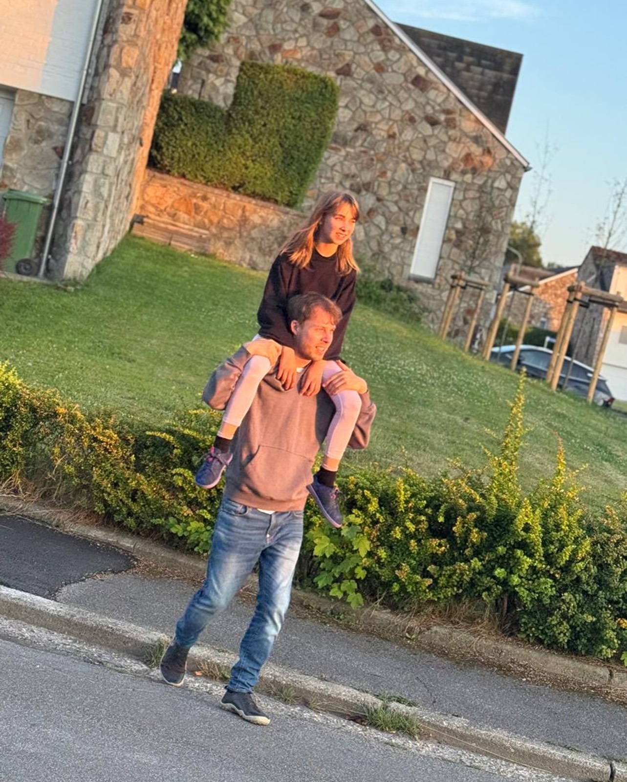 Heinz carrying Aylin on his shoulders, evening light, a street in Belgium.
